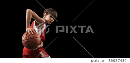 Cropped studio shot of boy, basketball player in uniform posing with ball in action isolated over black background. Flyer 86027482