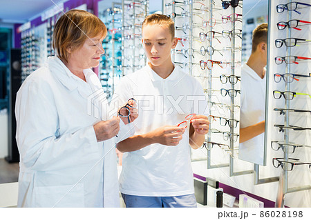 Professional mature female ophthalmologist helping teen boy to choose glasses in optical stor 86028198