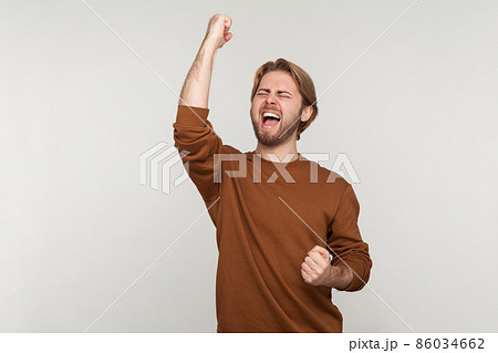 Portrait of excited man with beard wearing sweatshirt, expressing winning gesture with raised fists and screaming, celebrating victory. Indoor studio shot isolated on gray background. Portrait of excited man with beard wearing sweatshirt, expressing winning gesture with raised fists and screaming, celebrating victory. Indoor studio shot isolated on gray background. 86034662