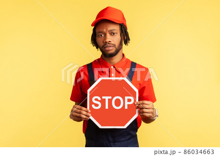 Portrait of handyman wearing blue overalls red hat and t-shirt holding red stop sign, prohibitions, looking at camera with serious expression. Indoor studio shot isolated on yellow background. 86034663