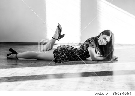 Winsome female lying on floor, looking at camera with smile and flirting expression, wearing dress and sandals. Black and white photography, indoor studio shot illuminated by sunlight from window. 86034664