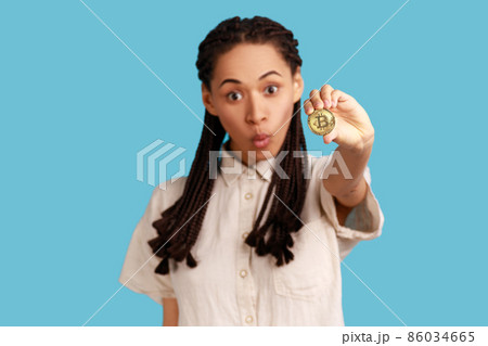 Portrait of excited astonished woman with black dreadlocks holding out bitcoin, coin of crypto currency, ecommerce, wearing white shirt. Indoor studio shot isolated on blue background. 86034665