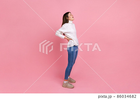 Full length portrait of girl touches injured back, suffers pain in lower lumbar muscular, arthritis, wearing white casual style sweater. Indoor studio shot isolated on pink background. 86034682