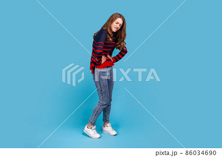 Full length portrait of woman wearing striped casual style sweater, frowning face from pain, suffering from painful period, gastritis or constipation. Indoor studio shot isolated on blue background. Full length portrait of woman wearing striped casual style sweater, frowning face from pain, suffering from painful period, gastritis or constipation. Indoor studio shot isolated on blue background. 86034690