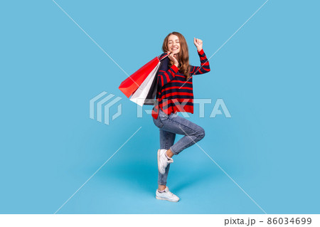 Full length portrait of positive woman wearing striped casual style sweater, standing with shopping bags, dancing, satisfied with her purchase. Indoor studio shot isolated on blue background. 86034699