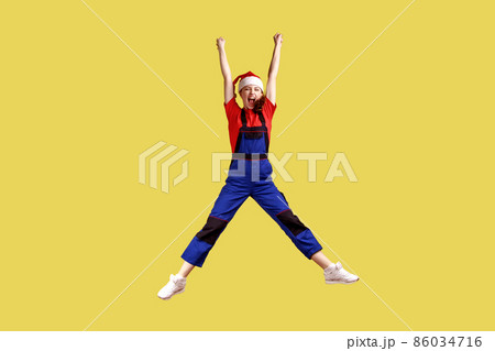 Full length portrait of excited happy female worker jumping high, finishing work before Christmas, wearing blue overalls and santa claus hat. Indoor studio shot isolated on yellow background. Full length portrait of excited happy female worker jumping high, finishing work before Christmas, wearing blue overalls and santa claus hat. Indoor studio shot isolated on yellow background. 86034716