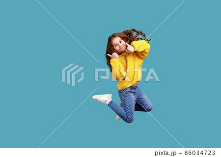 Full length portrait of happy charming little girl jumping up high and showing thumb up to camera, wearing yellow casual style sweater. Indoor studio shot isolated on blue background. Full length portrait of happy charming little girl jumping up high and showing thumb up to camera, wearing yellow casual style sweater. Indoor studio shot isolated on blue background. 86034723
