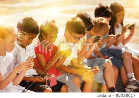 Smiling children sitting at urban street with mobile devices 86038017