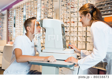 Teenage boy receiving eye examination on modern refractometer in optical store with professional female ophthalmologist 86038873