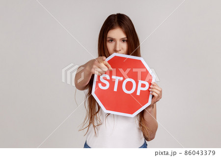 Serious woman holding big red stop road sign and pointing finger on you looking at camera with strict expression, warning, wearing white T-shirt. Indoor studio shot isolated on gray background. Serious woman holding big red stop road sign and pointing finger on you looking at camera with strict expression, warning, wearing white T-shirt. Indoor studio shot isolated on gray background. 86043379