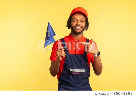 Happy handyman in uniform with dreadlocks pointing finger at Europe union flag in his hand and looking at camera with positive expression. Indoor studio shot isolated on yellow background. Happy handyman in uniform with dreadlocks pointing finger at Europe union flag in his hand and looking at camera with positive expression. Indoor studio shot isolated on yellow background. 86043395