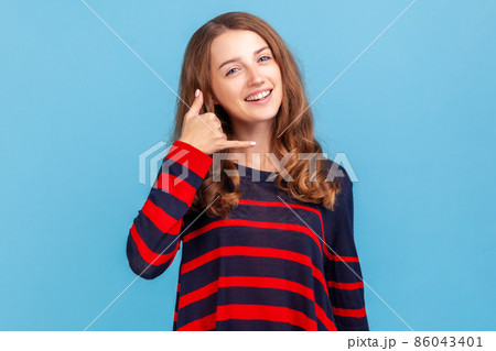 Hey handsome, call me. Smiling woman wearing striped casual style sweater flirting holding fingers near ear showing call gesture, answering call. Indoor studio shot isolated on blue background. Hey handsome, call me. Smiling woman wearing striped casual style sweater flirting holding fingers near ear showing call gesture, answering call. Indoor studio shot isolated on blue background. 86043401