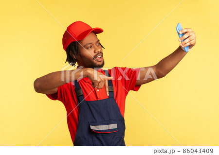Handyman wearing blue uniform having video call with client, pointing down, asking to subscribe on social network of his service industry. Indoor studio shot isolated on yellow background. Handyman wearing blue uniform having video call with client, pointing down, asking to subscribe on social network of his service industry. Indoor studio shot isolated on yellow background. 86043409
