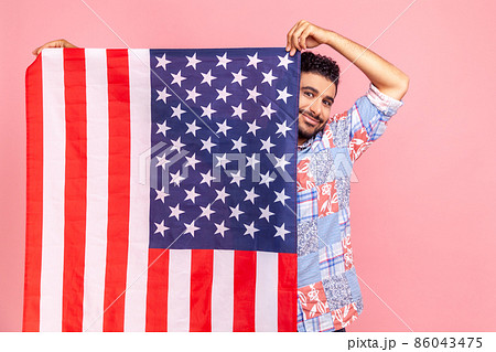 Portrait of attractive man holding USA flag in hands, looking at camera with toothy smile, proud of his social security in country, feeling in safe. Indoor studio shot isolated on pink background. 86043475