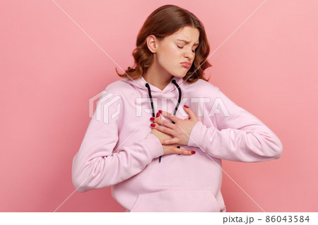 Portrait of curly haired teen girl in hoodie pressing on chest with painful expression, suffering sudden ache, heart attack, cardiac problems. Indoor studio shot isolated on pink background 86043584