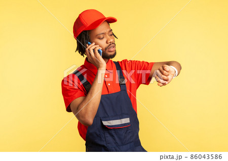 Portrait of concentrated courier wearing blue overalls talking phone with client, looking at smart watch, delivery service on time. Indoor studio shot isolated on yellow background. 86043586
