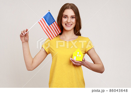 Portrait of happy teenager with brown hair holding flag of united states of america and paper house, dreaming about living in usa, green card. Indoor studio shot isolated on gray background. Portrait of happy teenager with brown hair holding flag of united states of america and paper house, dreaming about living in usa, green card. Indoor studio shot isolated on gray background. 86043648