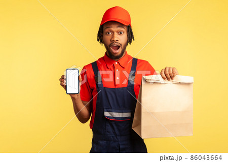 Wow, delivery service app. Deliveryman showing paper parcel and cellphone with mockup display for advertisement, looking shocked at camera. Indoor studio shot isolated on yellow background. 86043664