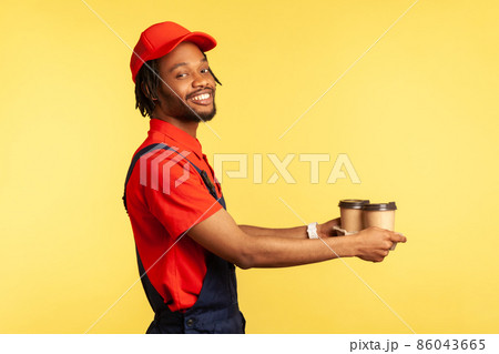 Side view portrait of smiling positive courier man wearing red T-shirt and blue uniform, giving take away coffee to client, delivery order. Indoor studio shot isolated on yellow background. 86043665