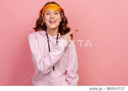 Portrait of happy curly haired teenage girl in hoodie and yellow sun visor smiling and pointing at empty space background, place for advertisement. Indoor studio shot isolated on pink background 86043727