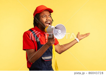 Attractive bearded handyman wearing red T-shirt and blue overalls holding megaphone, screaming, announcing discounts for service industry. Indoor studio shot isolated on yellow background. 86043728