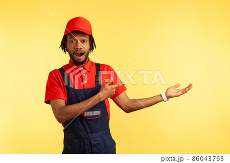 Astonished builder or courier wearing overalls, presenting copy space for advertisement, pointing finger aside, ad of delivery service. Indoor studio shot isolated on yellow background. 86043763