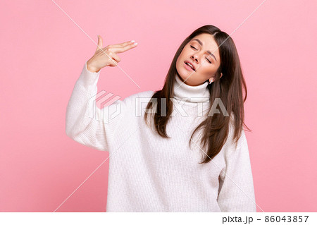 Unhappy woman stands with finger gun pointed to head, bored or tired, difficult to cope with problem, wearing white casual style sweater. Indoor studio shot isolated on pink background. 86043857