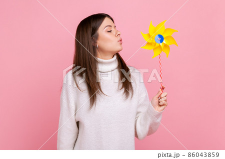 Portrait of carefree brunette woman blowing at paper windmill, playing with pinwheel toy on stick, wearing white casual style sweater. Indoor studio shot isolated on pink background. 86043859