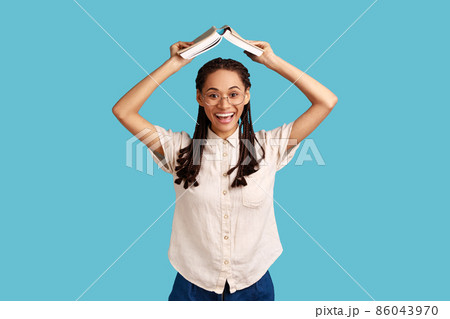 Portrait of positive woman with black dreadlocks and eyeglasses, standing holding book over head, studying and education, wearing white shirt. Indoor studio shot isolated on blue background. Portrait of positive woman with black dreadlocks and eyeglasses, standing holding book over head, studying and education, wearing white shirt. Indoor studio shot isolated on blue background. 86043970