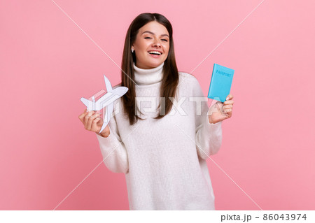 Portrait of excited female with passport and paper plane in hands rejoices future tour, looking at camera., wearing white casual style sweater. Indoor studio shot isolated on pink background. 86043974