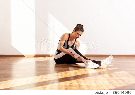 Woman shouting from pain in calf, touching injured leg, hurting to move sprained ligaments, wearing black sports top and tights. Full length studio shot illuminated by sunlight from window. Woman shouting from pain in calf, touching injured leg, hurting to move sprained ligaments, wearing black sports top and tights. Full length studio shot illuminated by sunlight from window. 86044090