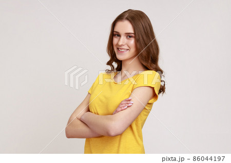 Portrait of positive young woman with brown wavy hair in yellow T-shirt standing with crossed arms, looking at camera with kindness and charming smile. Indoor studio shot isolated on gray background. 86044197