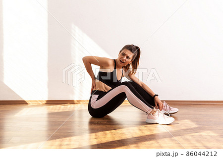 Athletic female frowning from pain in lower back, muscle strains and ligament sprains problem, wearing black sports top and tights. Full length studio shot illuminated by sunlight from window. Athletic female frowning from pain in lower back, muscle strains and ligament sprains problem, wearing black sports top and tights. Full length studio shot illuminated by sunlight from window. 86044212