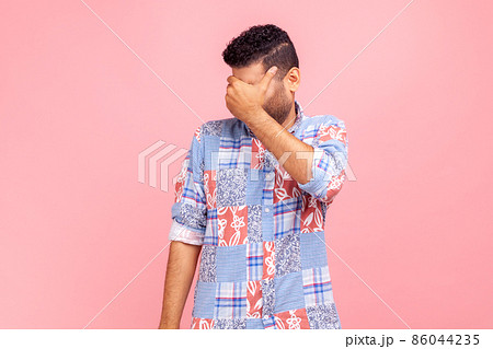 Don't want to look at this. Portrait of dark haired man in casual blue shirt covering eyes and turning with disgust from something shameful. Indoor studio shot isolated on pink background. 86044235