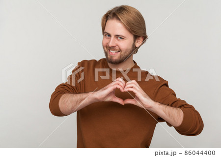 Portrait of happy attractive man with beard wearing sweatshirt, standing with heart or love gesture and looking at camera with toothy smile. Indoor studio shot isolated on gray background. 86044400