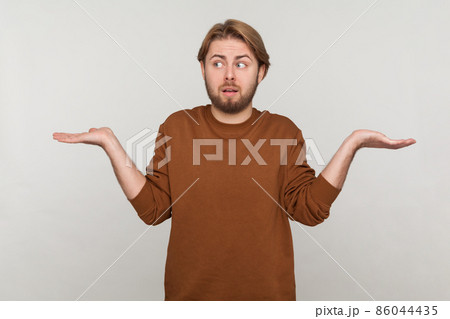 Portrait of man with beard wearing sweatshirt, shrugging shoulders as doesn't know answer, cant make decision, being uncertain, not sure. Indoor studio shot isolated on gray background. 86044435