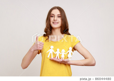 Portrait of happy satisfied female of young age holding paper family chain and showing thumb up, looking at camera with toothy smile. Indoor studio shot isolated on gray background. Portrait of happy satisfied female of young age holding paper family chain and showing thumb up, looking at camera with toothy smile. Indoor studio shot isolated on gray background. 86044480