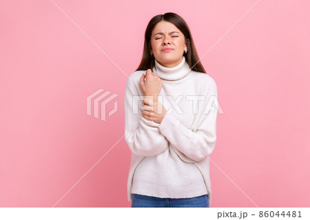 Upset woman standing with grimace of pain, massaging sore wrist, suffering hand injury or sprain, wearing white casual style sweater. Indoor studio shot isolated on pink background. 86044481