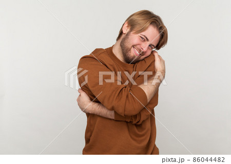 Portrait of happy handsome man with beard wearing sweatshirt, standing and hugging himself with toothy smile enjoying, positive self-esteem. Indoor studio shot isolated on gray background. Portrait of happy handsome man with beard wearing sweatshirt, standing and hugging himself with toothy smile enjoying, positive self-esteem. Indoor studio shot isolated on gray background. 86044482