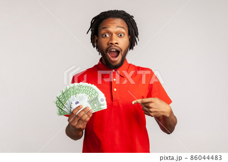 Astonished man with open mouth wearing red casual style T-shirt, hold lot of money, pointing at euro banknotes, shocked by sudden winning. Indoor studio shot isolated on gray background. Astonished man with open mouth wearing red casual style T-shirt, hold lot of money, pointing at euro banknotes, shocked by sudden winning. Indoor studio shot isolated on gray background. 86044483