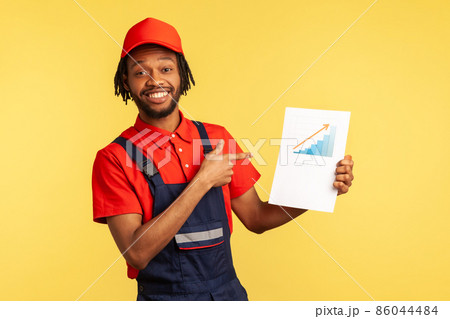Portrait of smiling satisfied workman with beard wearing blue uniform, holding paper with graph showing financial increase of service industry. Indoor studio shot isolated on yellow background. Portrait of smiling satisfied workman with beard wearing blue uniform, holding paper with graph showing financial increase of service industry. Indoor studio shot isolated on yellow background. 86044484
