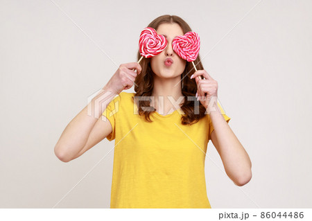 Portrait of childish woman of young age in urban style T-shirt covering eye with lollipops, making kissing gesture to camera, playing with sweet candy. Indoor studio shot isolated on gray background. Portrait of childish woman of young age in urban style T-shirt covering eye with lollipops, making kissing gesture to camera, playing with sweet candy. Indoor studio shot isolated on gray background. 86044486