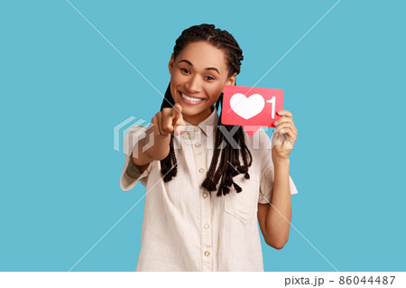 Portrait of happy positive woman with black dreadlocks showing blogger heart icon and pointing to camera, asking to click button, wearing white shirt. Indoor studio shot isolated on blue background. Portrait of happy positive woman with black dreadlocks showing blogger heart icon and pointing to camera, asking to click button, wearing white shirt. Indoor studio shot isolated on blue background. 86044487