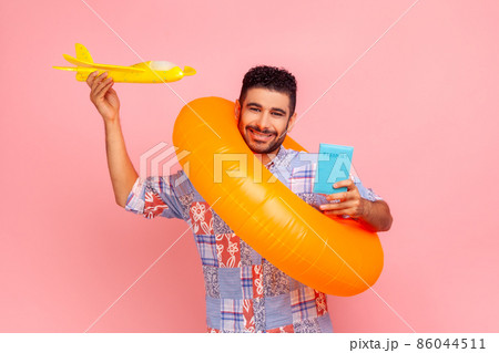happy satisfied tourist man in blue shirt standing with rubber ring, holding passport document and airplane mockup, enjoying travel tour. Indoor studio shot isolated on pink background. happy satisfied tourist man in blue shirt standing with rubber ring, holding passport document and airplane mockup, enjoying travel tour. Indoor studio shot isolated on pink background. 86044511