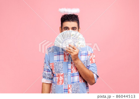 Angelic rich bearded man wearing blue casual style shirt with nimbus on head peeking out of dollar banknotes, rejoicing big profit earn. Indoor studio shot isolated on pink background. 86044513