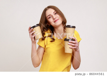 Delivery service. Friendly happy teenager woman of young age in yellow T-shirt holding coffee, drinks in disposable cups, looking at camera with smile. Indoor studio shot isolated on gray background. 86044561