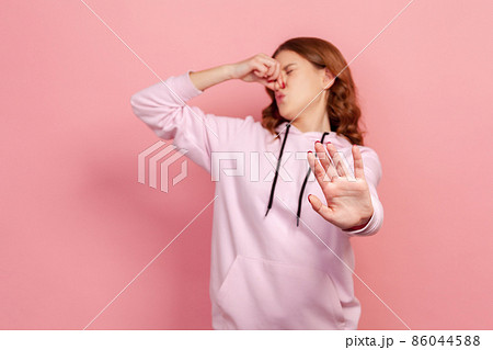 Portrait of displeased teen girl in hoodie grimacing in disgust and holding breath, grabbing nose and showing stop to avoid stinky odor, fart gases. Indoor studio shot isolated on pink background 86044588