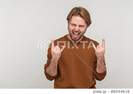 Portrait of man with beard wearing sweatshirt, showing rock and roll sign hand gesture, screaming, having excited facial expression. Indoor studio shot isolated on gray background. 86044616