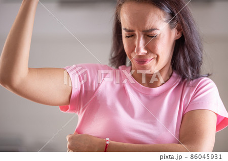 Woman smells herself with repulsive facial expression and sweat patches on her pink shirt Woman smells herself with repulsive facial expression and sweat patches on her pink shirt 86045931