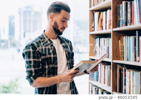 Young male librarian in casual clothing making order near bookshelves in college campus Young male librarian in casual clothing making order near bookshelves in college campus 86053595